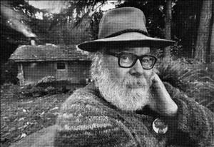 A man with a bushy white beard, eyeglasses, and a floppy hat looks into the camera. There is a small cabin behind him with smoke coming out of a stovepipe. He is in the forest. 